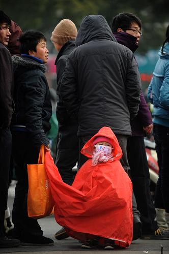 1月15日,来自安徽阜阳的1岁半小女孩王晓玉与母亲(左)在宁波一个火车票代售窗口前排队买票,为抵御寒风,母亲给她裹上了一件雨衣。当日,正值春运前铁路购票的高峰时期,大批旅客为顺利买到从宁波返乡的火车票,冒着凛冽的寒风在购票点排队等候,有人甚至早早地在预售日车票开售前一天的凌晨来到窗口前排队。新华社记者 黄宗治 摄