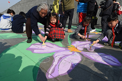 3月28日，南京大屠杀幸存者伍秀英（前左一）与小学生志愿者一起用美术颜料手绘紫金草图案。当日，“紫金草手绘行动”在侵华日军南京大屠杀遇难同胞纪念馆雕塑广场举行，南京艺术学院的画家和来自社会各界的200名志愿者一起，用美术颜料手绘紫金草图案，将和平之花“种植”在纪念馆雕塑广场的地面上。
